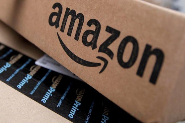 Caja de cartón de Amazon en primer plano, con el logotipo negro visible, sobre otro paquete con cinta adhesiva de Amazon Prime.
