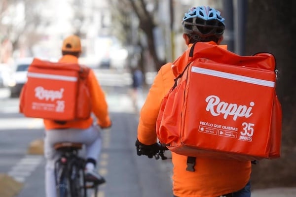 Dos repartidores en bicicleta, con uniformes naranjas y mochilas térmicas rojas de Rappi, circulan por una calle urbana desenfocada.
